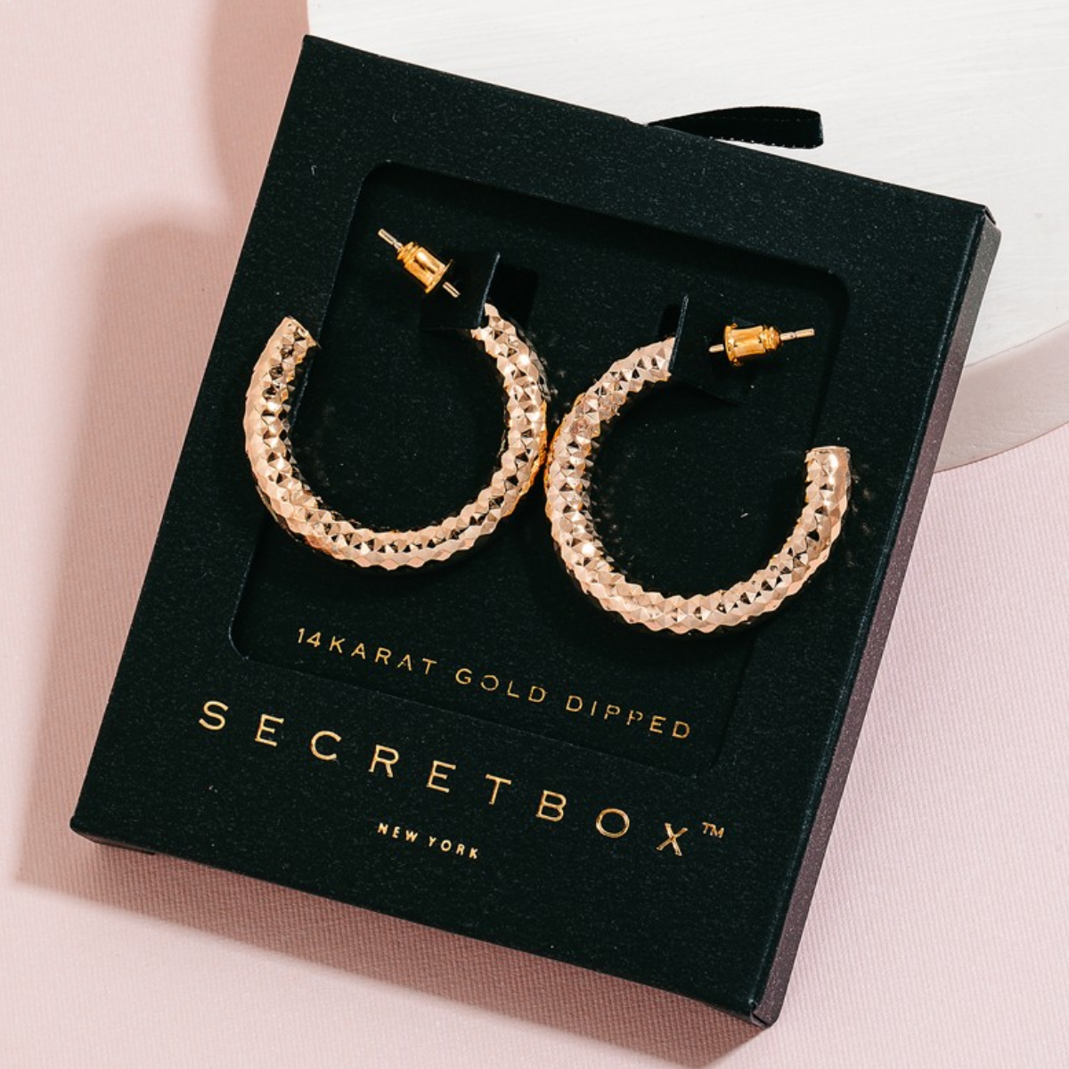 Pair of Secret Box Gold Dipped Textured Hoop Earrings in Gold by FASHION GO in a black box labeled "SECRET BOX NEW YORK," displayed against a pink background.