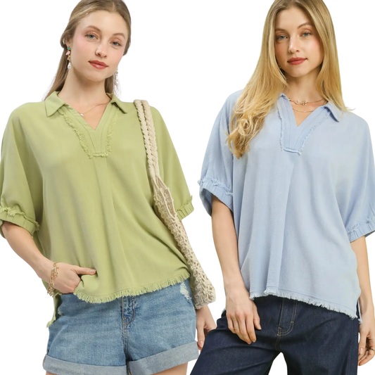 Two women wear the Umgee Linen Frayed Edge Short Sleeve Top—one in light green, one in light blue—with denim shorts. These loose-fit tops feature split necks and frayed edges.