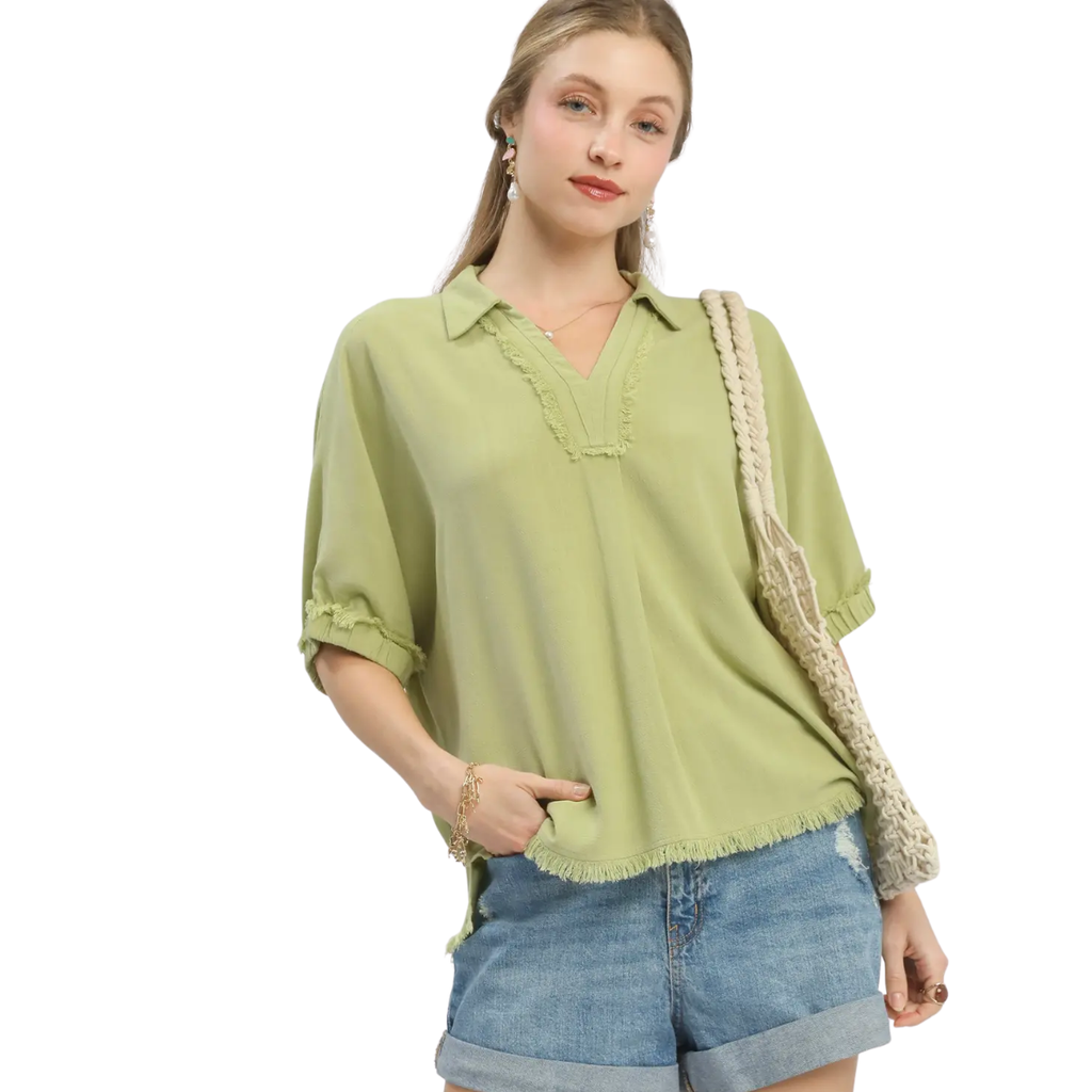 A woman models the Umgee Linen Frayed Edge Short Sleeve Top in light green, paired with denim shorts and a beige woven shoulder bag, standing against a white background.
