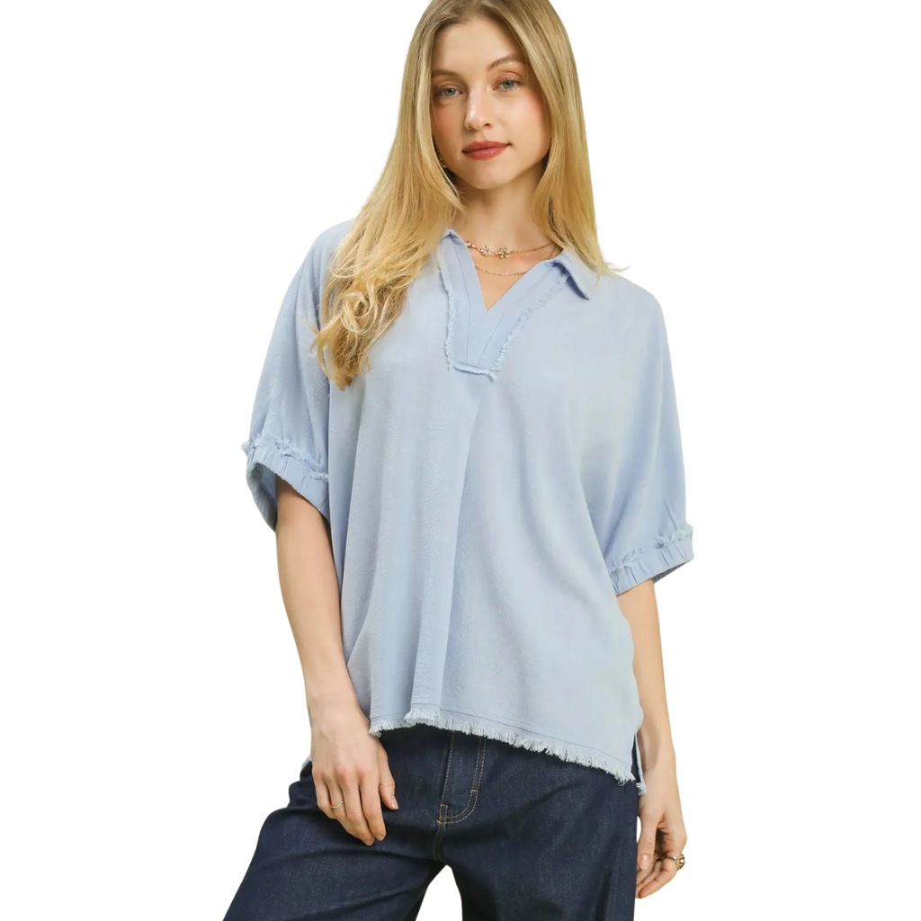 A woman wears the Umgee Linen Frayed Edge Short Sleeve Top in light blue with a split neck, paired with dark jeans, standing against a white background.