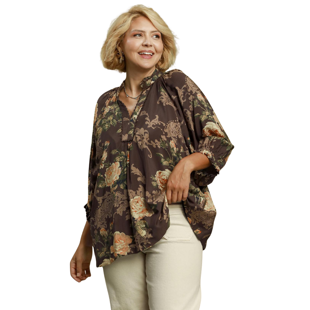 A woman wears an Umgee Floral Ruffle Neck Split V-Neck Blouse with a ruffle-trimmed collar and split V-neckline, paired with cream-colored pants.