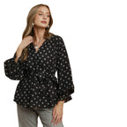 A woman models the Umgee Polka Dot Split Neck Blouse with Ruffle Cuffs, featuring long sleeves and a waist tie, paired with gray pants against a white background.
