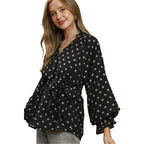 A woman is wearing the Umgee Polka Dot Split Neck Blouse with Ruffle Cuffs, featuring black fabric with white polka dots, long sleeves finished with ruffle cuffs, and a flattering waist tie, styled effortlessly with light-colored pants.