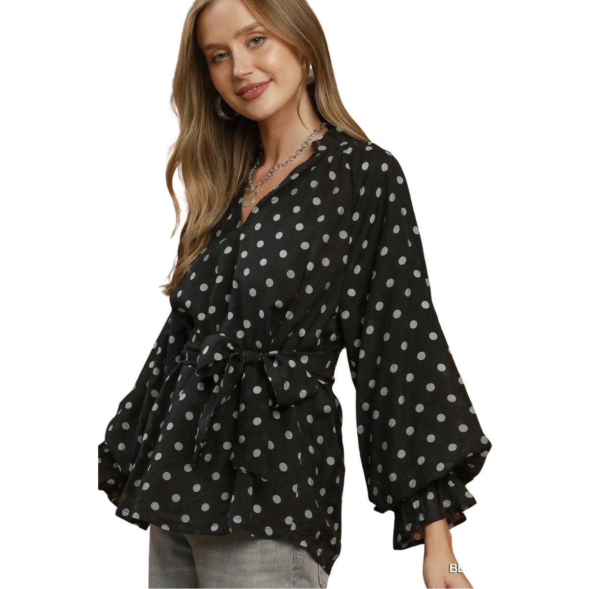 A woman is wearing the Umgee Polka Dot Split Neck Blouse with Ruffle Cuffs, featuring black fabric with white polka dots, long sleeves finished with ruffle cuffs, and a flattering waist tie, styled effortlessly with light-colored pants.