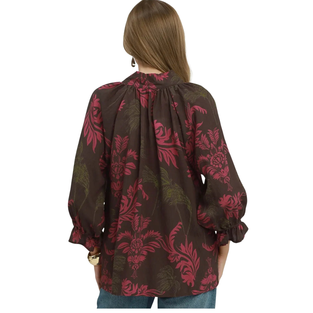 A woman with long brown hair is seen from the back wearing the Umgee Botanical Print Split Neck Blouse by Umgee, which features a loose fit, long sleeves, and a red and green floral pattern.