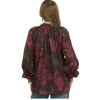 A woman with long brown hair is seen from the back wearing the Umgee Botanical Print Split Neck Blouse by Umgee, which features a loose fit, long sleeves, and a red and green floral pattern.
