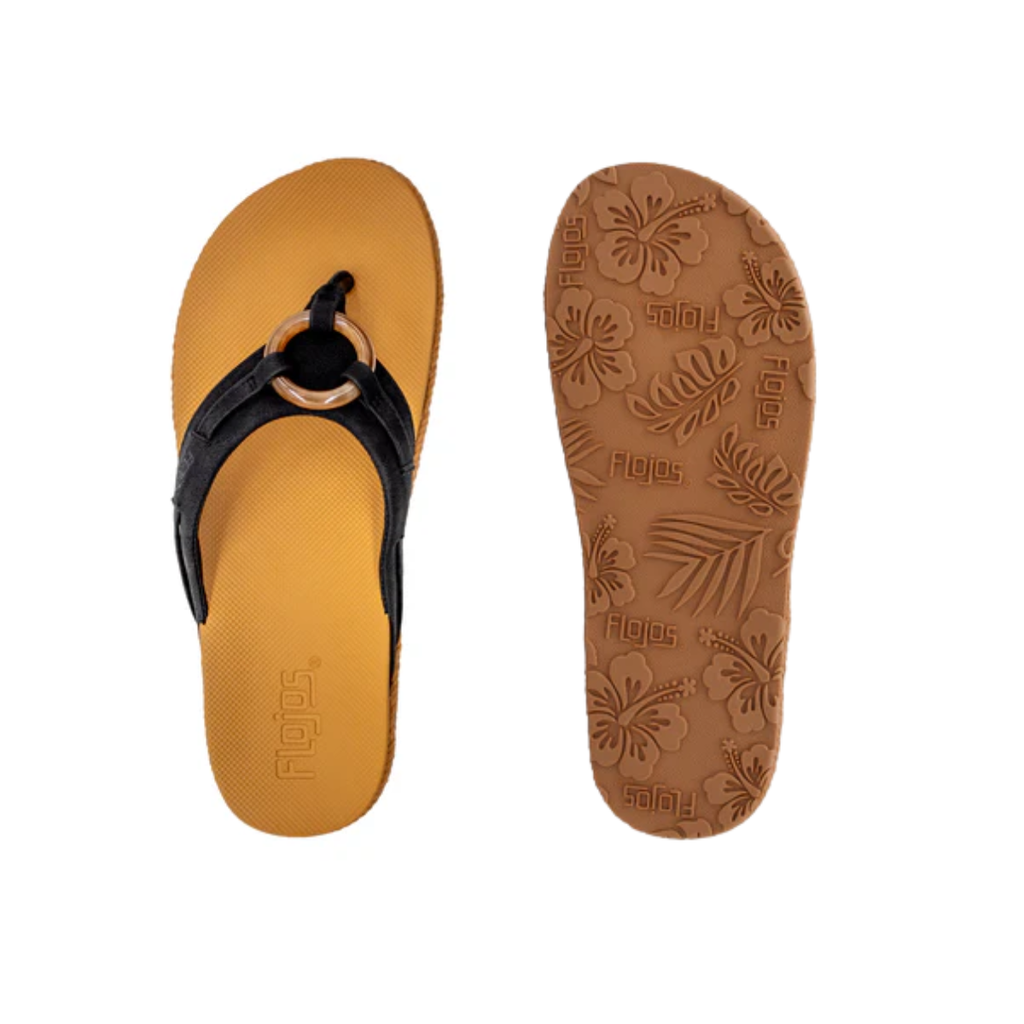 The FLOJOS Viernes Flip Flop is a women's sandal with tan soles, black faux leather straps, and a circular buckle. It features a memory foam footbed, floral-patterned sole for comfort and style, plus the distinctive FLOJOS branding.
