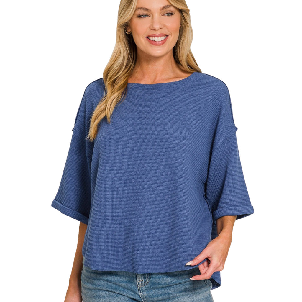 A woman stands smiling against a white background, wearing the Zenana Double Knit Exposed Seam Curved Hem Top in blue with denim shorts.