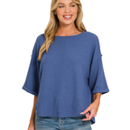 A woman stands smiling against a white background, wearing the Zenana Double Knit Exposed Seam Curved Hem Top in blue with denim shorts.