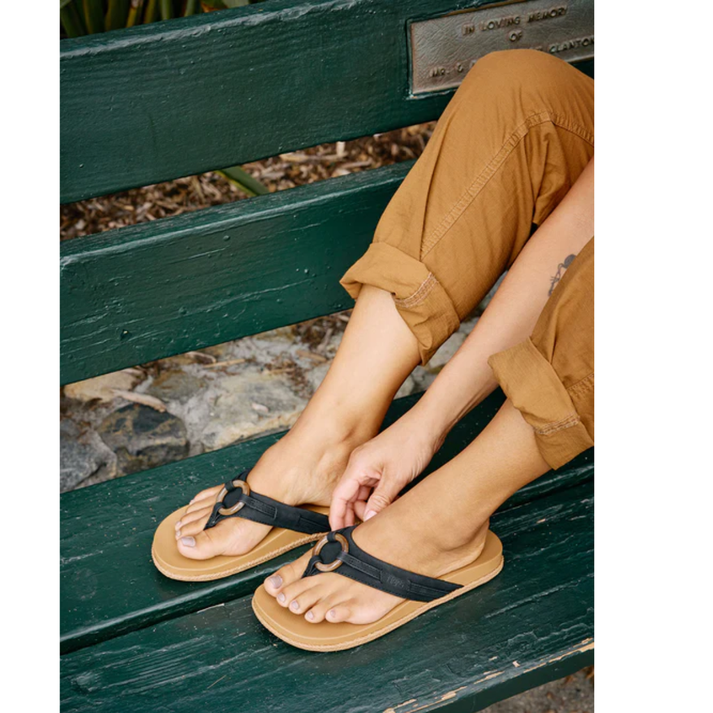 A person in tan pants sits on a green bench, adjusting FLOJOS Viernes Flip Flop women’s sandals with faux leather straps and a cushioned memory foam footbed on their feet.