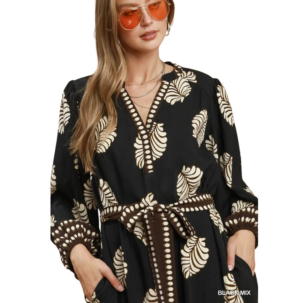 A woman wears orange sunglasses and the Umgee Leaf Print Dress—a black midi dress featuring beige leaf patterns, polka dot trim, a matching waist tie, and convenient pockets—for a stylish transitional look.