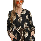 A woman wears orange sunglasses and the Umgee Leaf Print Dress—a black midi dress featuring beige leaf patterns, polka dot trim, a matching waist tie, and convenient pockets—for a stylish transitional look.
