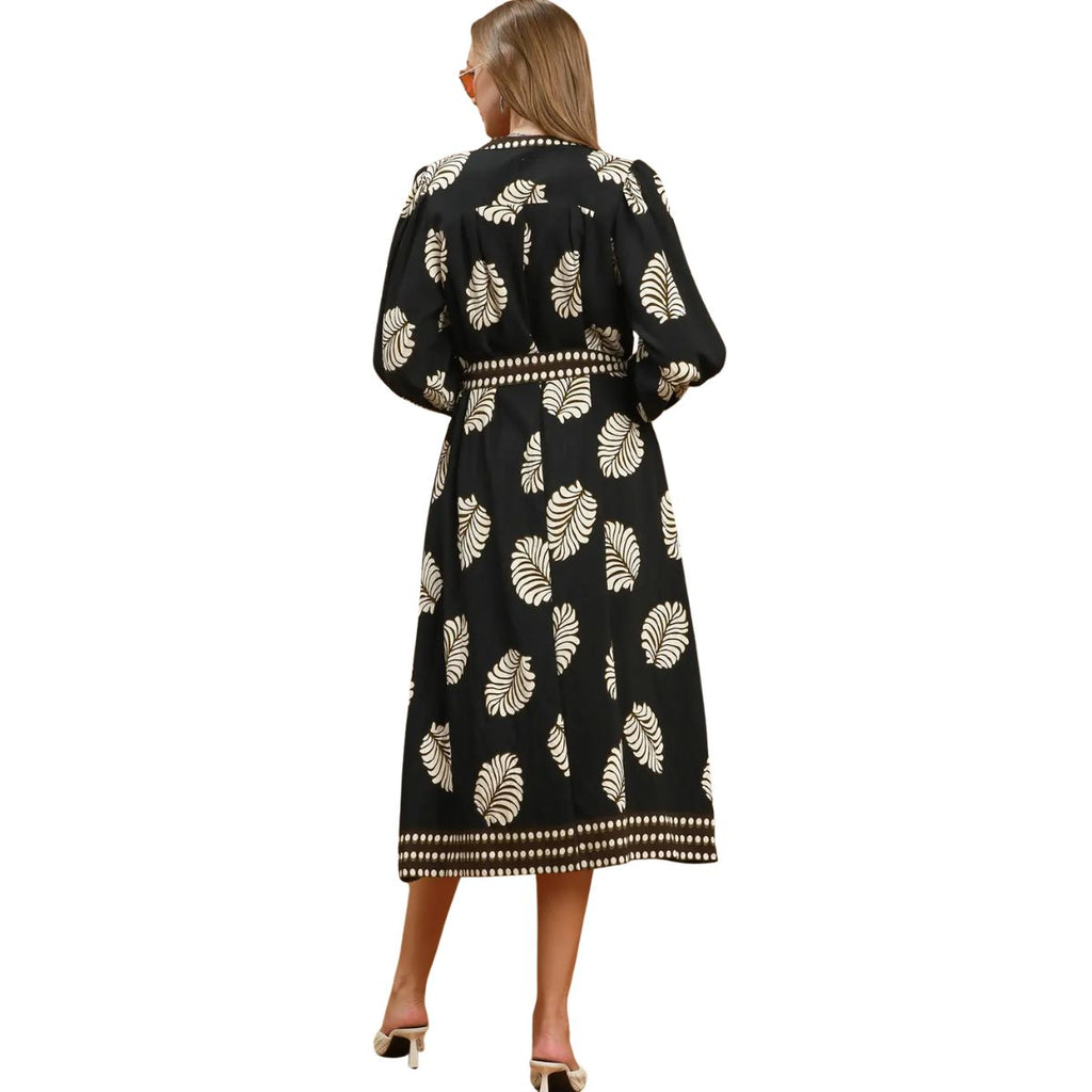 A woman stands with her back to the camera, wearing the Umgee Leaf Print Dress—a long black dress featuring white botanical leaf patterns and white trim—paired with white high-heeled shoes.