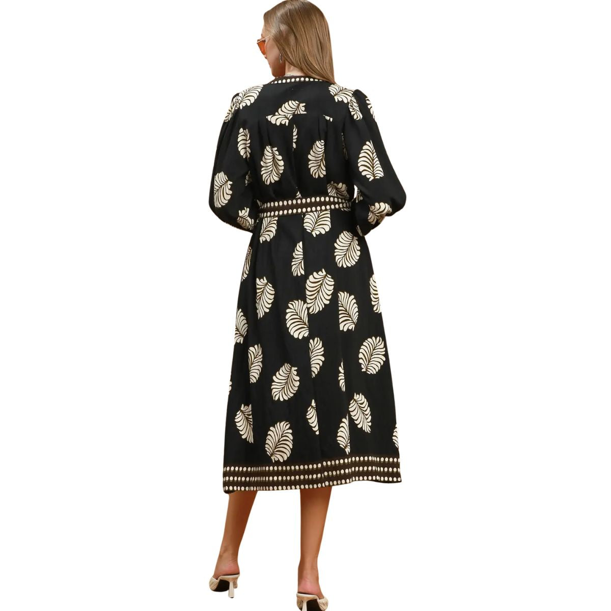 A woman stands with her back to the camera, wearing the Umgee Leaf Print Dress—a long black dress featuring white botanical leaf patterns and white trim—paired with white high-heeled shoes.