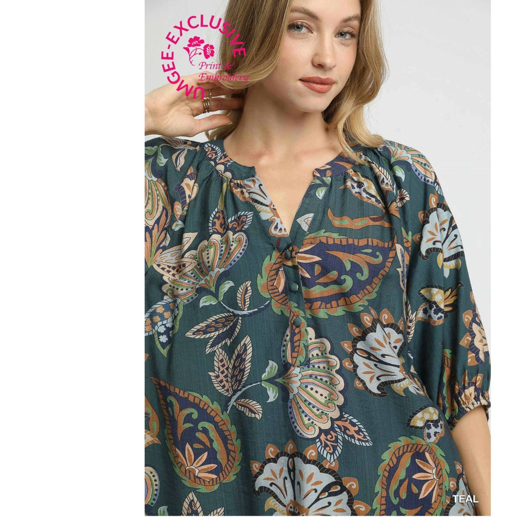 A woman wearing the Umgee Boho Paisley Puff Sleeve Blouse, featuring a teal floral pattern and three-quarter sleeves with a split neckline, stands against a plain background. A pink “VIGEE EXCLUSIVE” stamp appears in the top left corner.