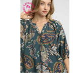 A woman wearing the Umgee Boho Paisley Puff Sleeve Blouse, featuring a teal floral pattern and three-quarter sleeves with a split neckline, stands against a plain background. A pink “VIGEE EXCLUSIVE” stamp appears in the top left corner.