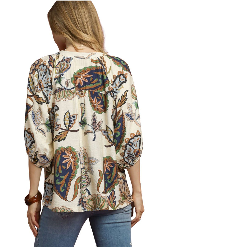 A woman stands with her back to the camera, wearing the Umgee Boho Paisley Puff Sleeve Blouse and blue jeans.