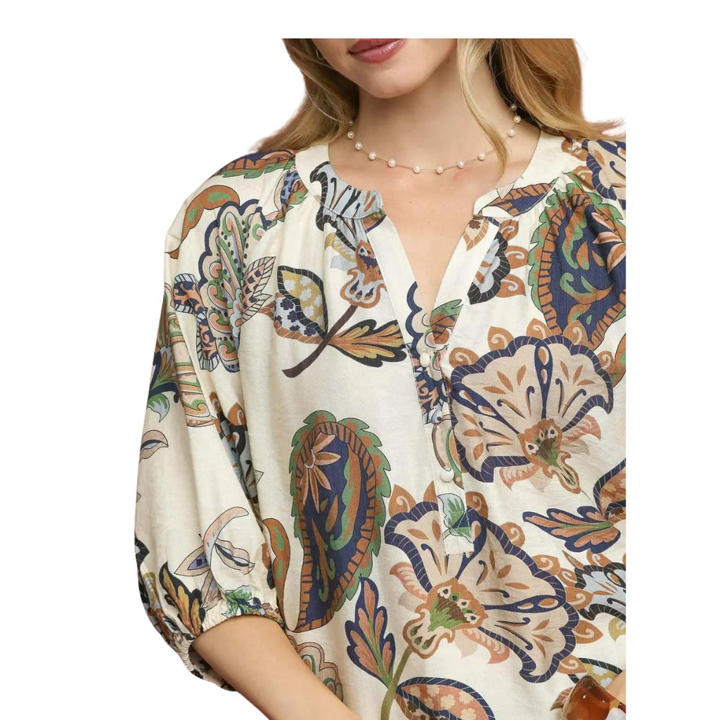 A woman wearing the Umgee Boho Paisley Puff Sleeve Blouse, featuring a cream hue with colorful floral and leaf print, three-quarter puff sleeves, a split round neckline, and styled with a delicate beaded necklace.