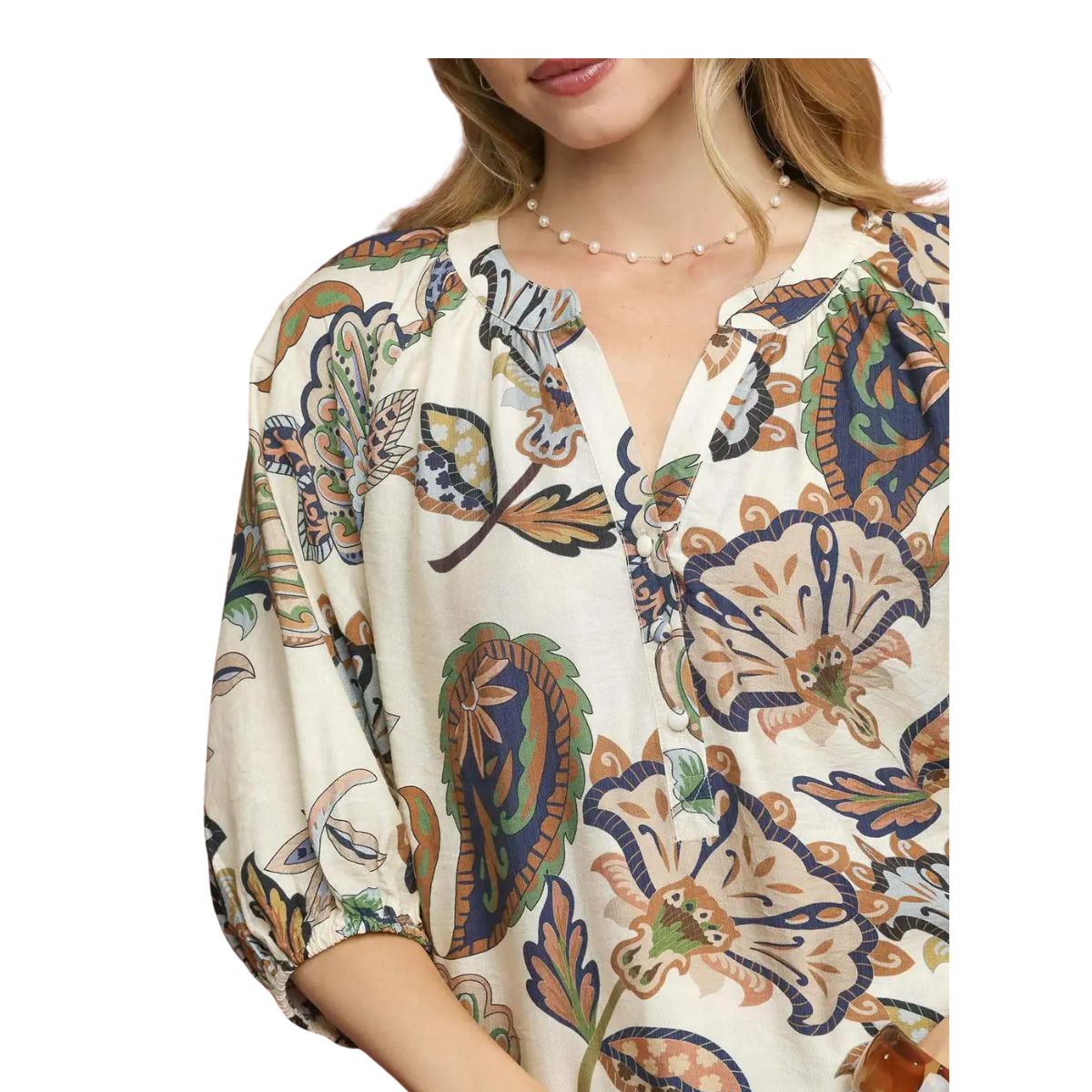 A woman wearing the Umgee Boho Paisley Puff Sleeve Blouse, featuring a cream hue with colorful floral and leaf print, three-quarter puff sleeves, a split round neckline, and styled with a delicate beaded necklace.
