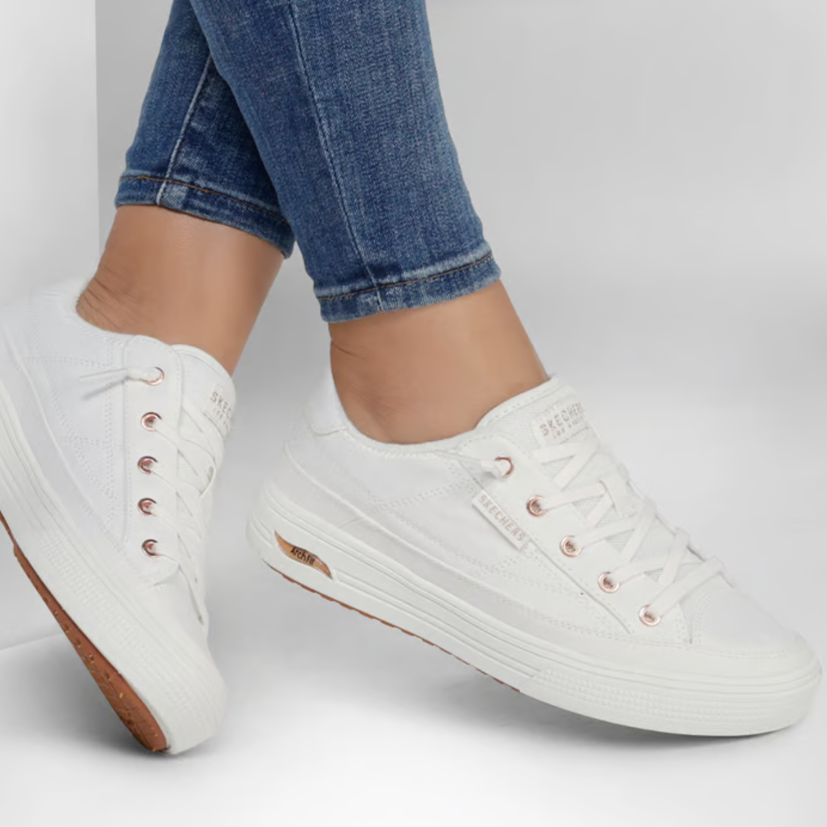 A person wearing cuffed blue jeans and SKECHERS USA INC Skechers Arch Fit Arcade White Sneaker - Meet Ya There, featuring arch support and rubber soles, stands against a white background.