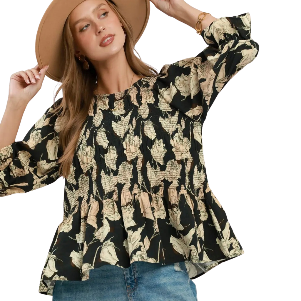 A woman wearing the Umgee Mix Floral Smocked Yoke Ruffle Sleeve Blouse and a tan hat with blue jeans poses, her hand adjusting the brim of her hat.