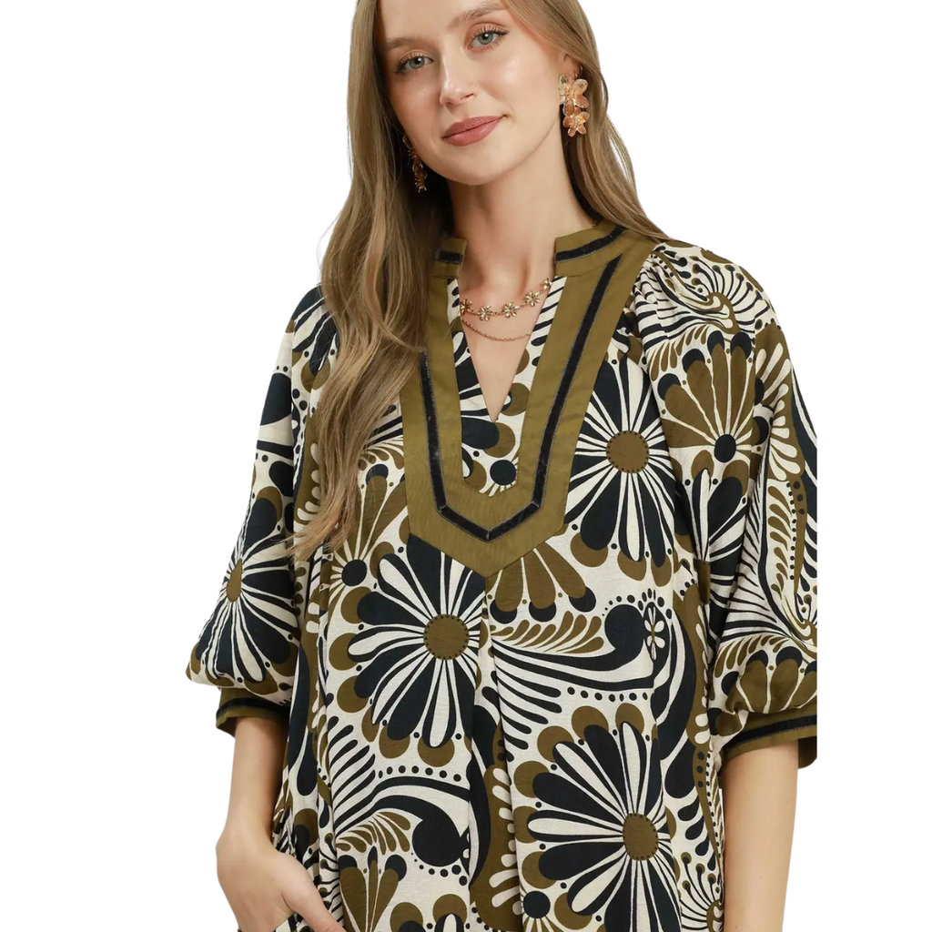 A woman wears the Umgee Abstract Print Midi Dress, featuring a flowy silhouette, abstract black, white, and olive green designs, and a V-neckline with decorative trim.