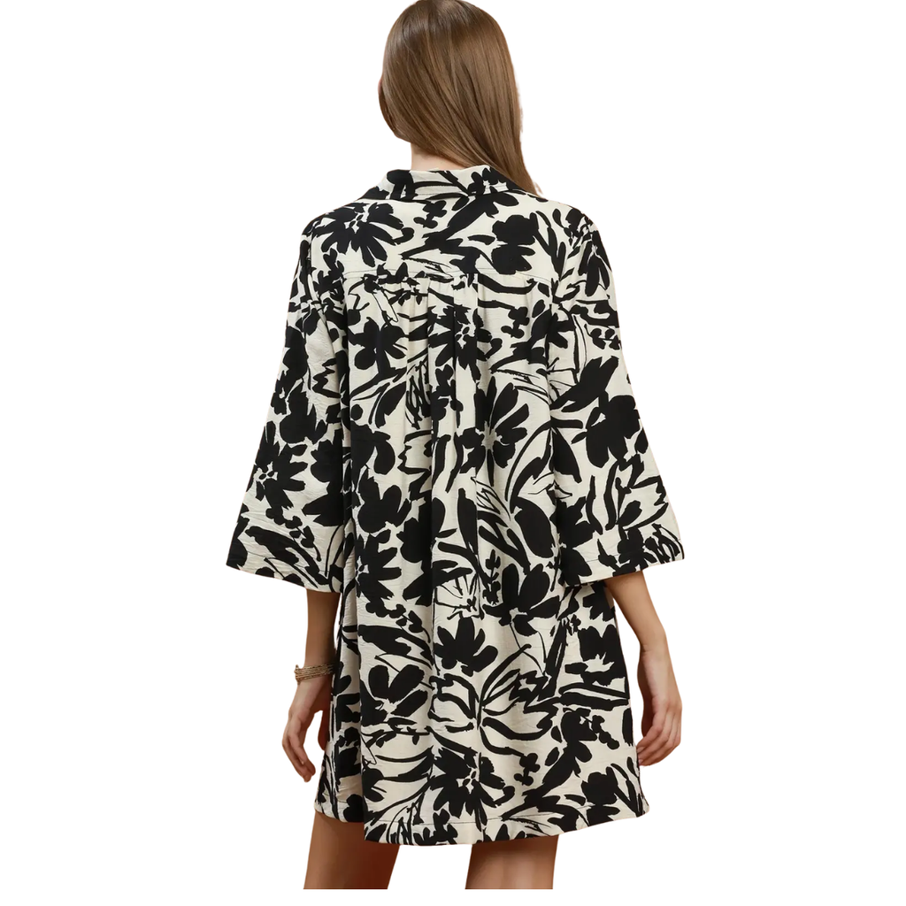 A woman with long hair is seen from behind wearing the Umgee Floral Print Split Neck Relaxed Shift Dress, featuring a loose fit and black floral print on white.