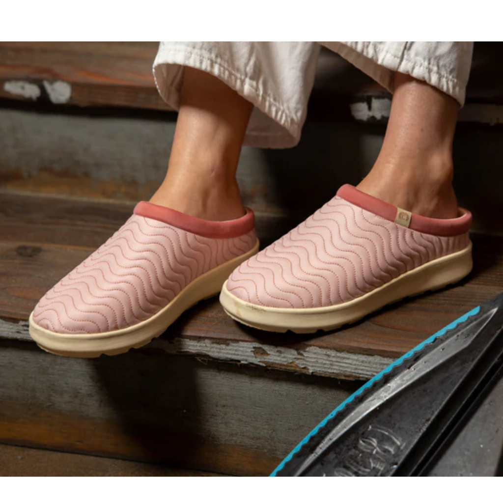 A person wearing COBIAN Nami Flip Slips in light pink, featuring wavy stitching and an EVA cupsole, stands on wooden steps. Only their lower legs and feet are visible, highlighting this closed-toe comfort footwear.