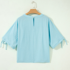 The Tie Half Sleeve Pleated Detail Babydoll Blouse by FASHION GO is shown in light blue on a hanger, viewed from the back. Made from lightweight, breathable fabric, it features a button closure at the neckline and tied accents on its half sleeves.