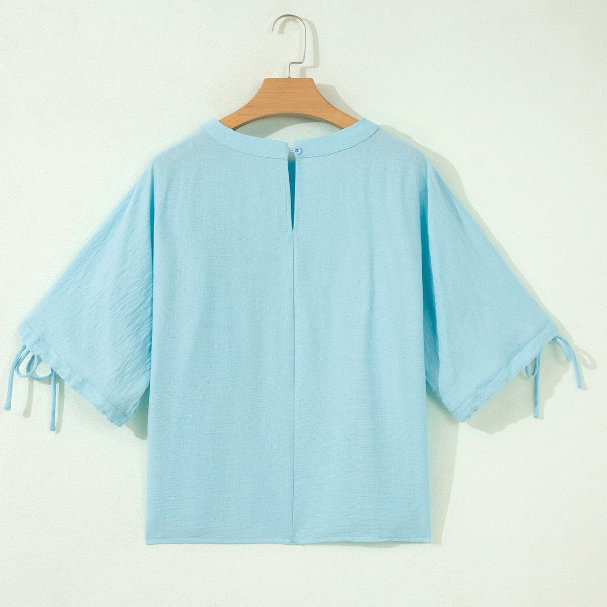 The Tie Half Sleeve Pleated Detail Babydoll Blouse by FASHION GO is shown in light blue on a hanger, viewed from the back. Made from lightweight, breathable fabric, it features a button closure at the neckline and tied accents on its half sleeves.