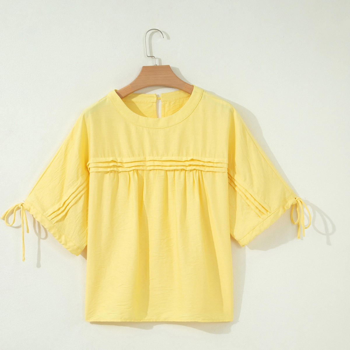 The FASHION GO Tie Half Sleeve Pleated Detail Babydoll Blouse is a light yellow, short-sleeve top featuring a round neckline, horizontal pleated accents on the chest, and tied ribbons on the sleeves. Crafted from a lightweight, breathable fabric, it is shown displayed on a wooden hanger against a white background.