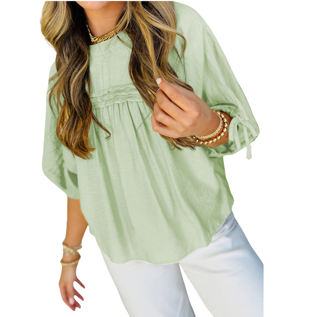 A woman is shown from shoulders to knees against a white background, wearing the FASHION GO Tie Half Sleeve Pleated Detail Babydoll Blouse in light green—crafted from lightweight, breathable fabric with half sleeves and pleated details—paired with white pants and gold jewelry.