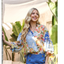 A woman with long blonde hair stands outdoors in the FASHION GO Floral Print Button Down Top and blue high-waisted pants with gold buttons, framed by large green plants in the background.