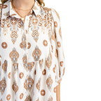 A person is shown from the waist up, wearing the Gigio Printed Shirt Dress featuring long sleeves, a collared neckline, and a brown and orange floral paisley print, styled over a pastel blue dress against a white background.