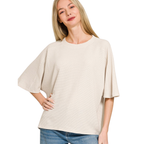 A woman wears a Zenana Double Knit Short Sleeve Top in light beige with blue jeans, standing with one hand on her hip and smiling against a white background.