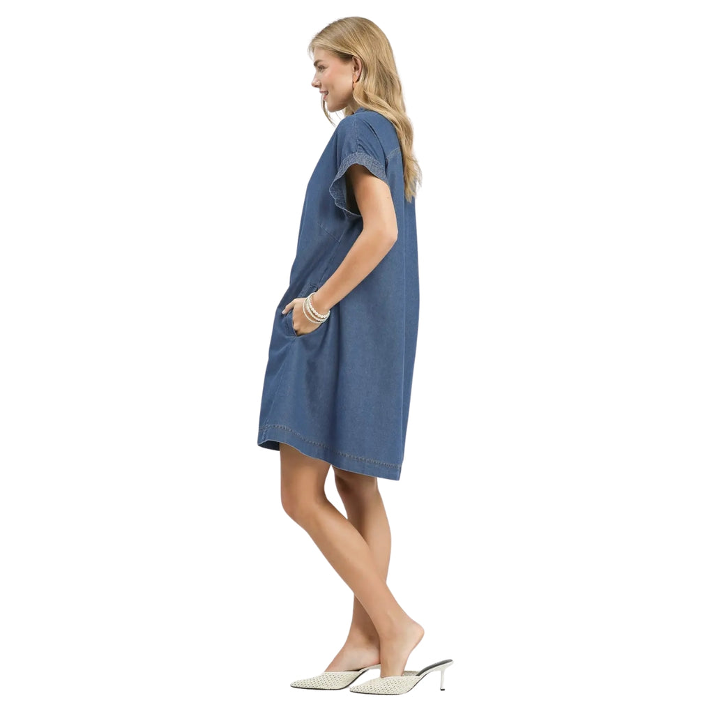 A woman wears the Umgee Chambray Button-Front Shirt Dress with white heeled shoes, standing in profile with hands in pockets against a white background.
