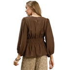 A woman is shown from the back, wearing the Umgee Smocked Waist Long Sleeve Peplum Blouse in brown, featuring a subtle peplum hem, paired with light patterned pants, against a white background.