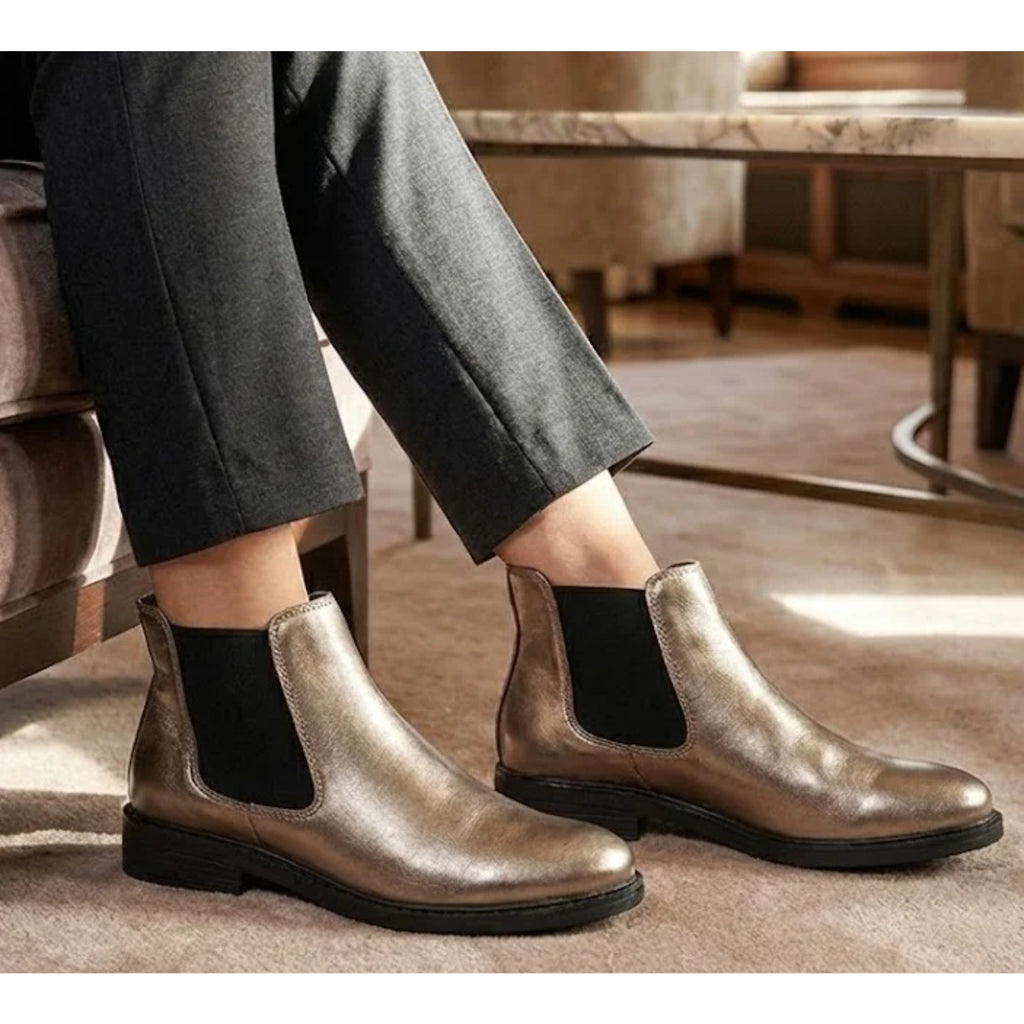 Wearing the Tiffany Chelsea Boot by Eric Michael in metallic pewter with black elastic side panels, a person sits indoors on a beige carpet next to a table and armchair.