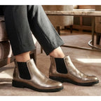 Wearing the Tiffany Chelsea Boot by Eric Michael in metallic pewter with black elastic side panels, a person sits indoors on a beige carpet next to a table and armchair.