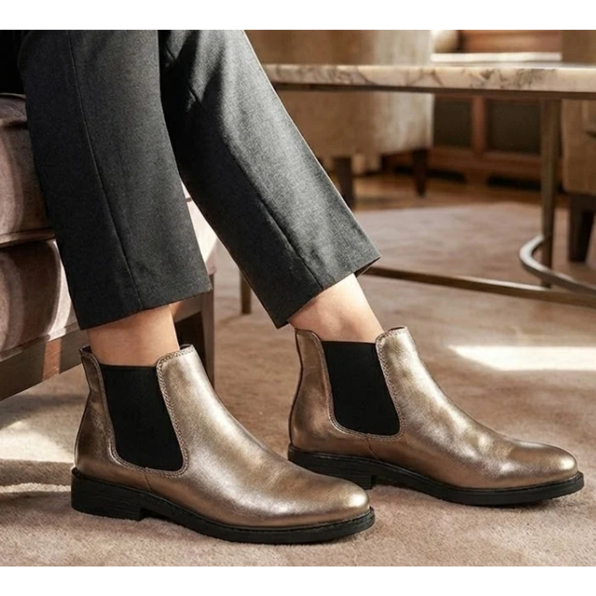 Wearing the Tiffany Chelsea Boot by Eric Michael in metallic pewter with black elastic side panels, a person sits indoors on a beige carpet next to a table and armchair.