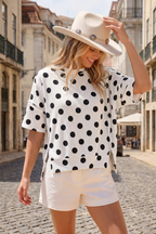 A woman in a white hat, Celeste Clothing’s Women’s Polka Dot Rib Knit Top, and white shorts stands on a cobblestone street lined with buildings, looking down and smiling.