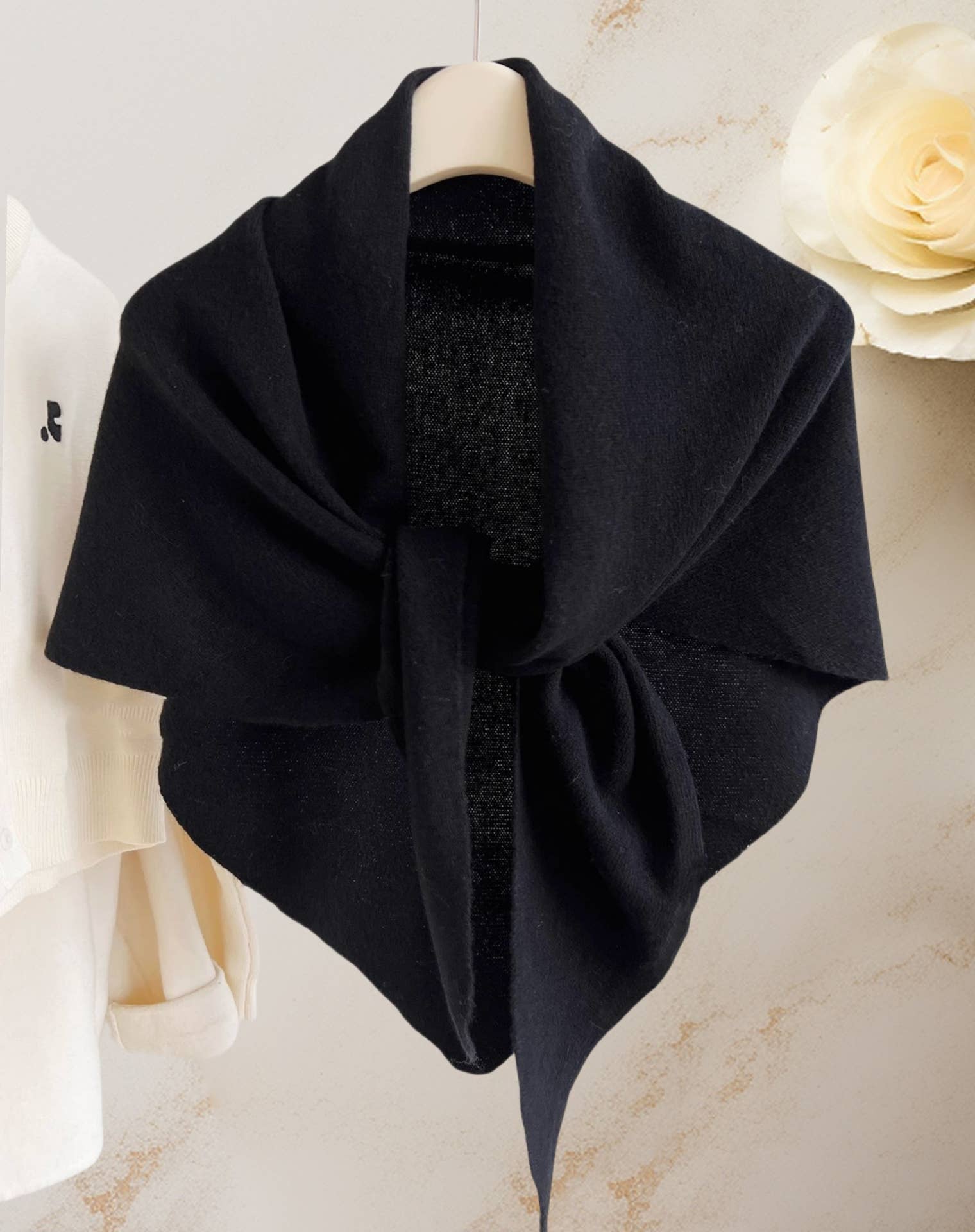 The JC Sunny Fashion Cashmere-Blend Knit Triangle Shawl with Cross-Front Design is draped over a hanger against a light background, with part of a white sweater and decorative flower visible at the edges.