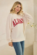 A woman wears an oversized cream-colored ALABAMA Cloudy Soft Knit Long Sleeve Top with red lettering across the chest and blue jeans.