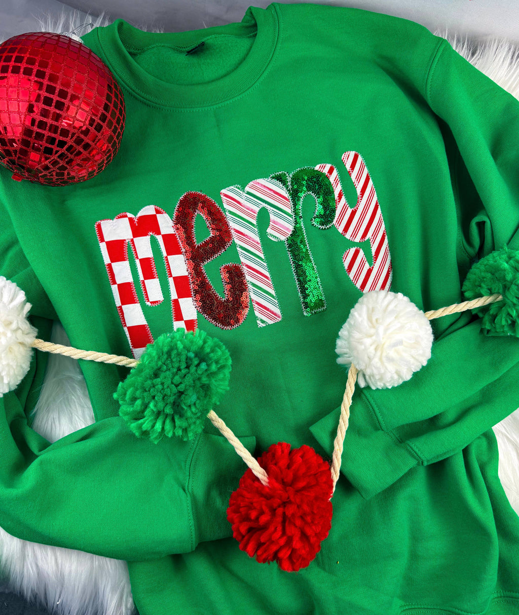Green Merry Sequin Christmas Sweatshirt featuring the word “merry” in patterned, sequined letters, decorated with a pom-pom garland and a red ornament.
