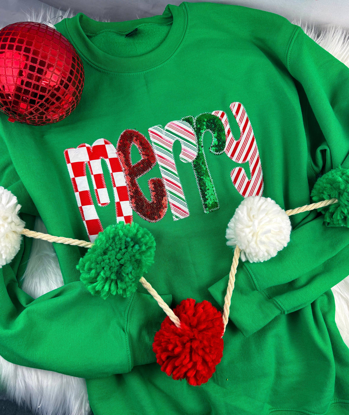 Green Merry Sequin Christmas Sweatshirt featuring the word “merry” in patterned, sequined letters, decorated with a pom-pom garland and a red ornament.