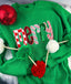 Green Merry Sequin Christmas Sweatshirt featuring the word “merry” in patterned, sequined letters, decorated with a pom-pom garland and a red ornament.