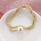 The Alabama Gold & Pearl Logo Stretch Bracelet Set by Seasons Jewelry features gold beaded stretch bracelets adorned with a white pearl and a red letter "A," echoing University of Alabama-inspired style, beautifully displayed on a white dish against a pink glittery background.