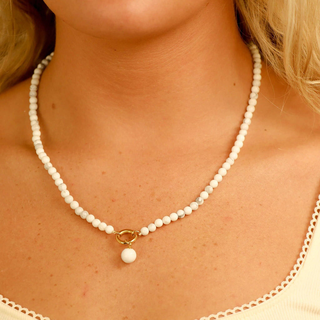 A close-up of a person wearing the WJW Semi Precious Natural Stone Charm Keeper Necklace, featuring a white beaded design with an 18K Gold PVD Coated clasp and a single round white pendant, shown against cream-colored clothing.