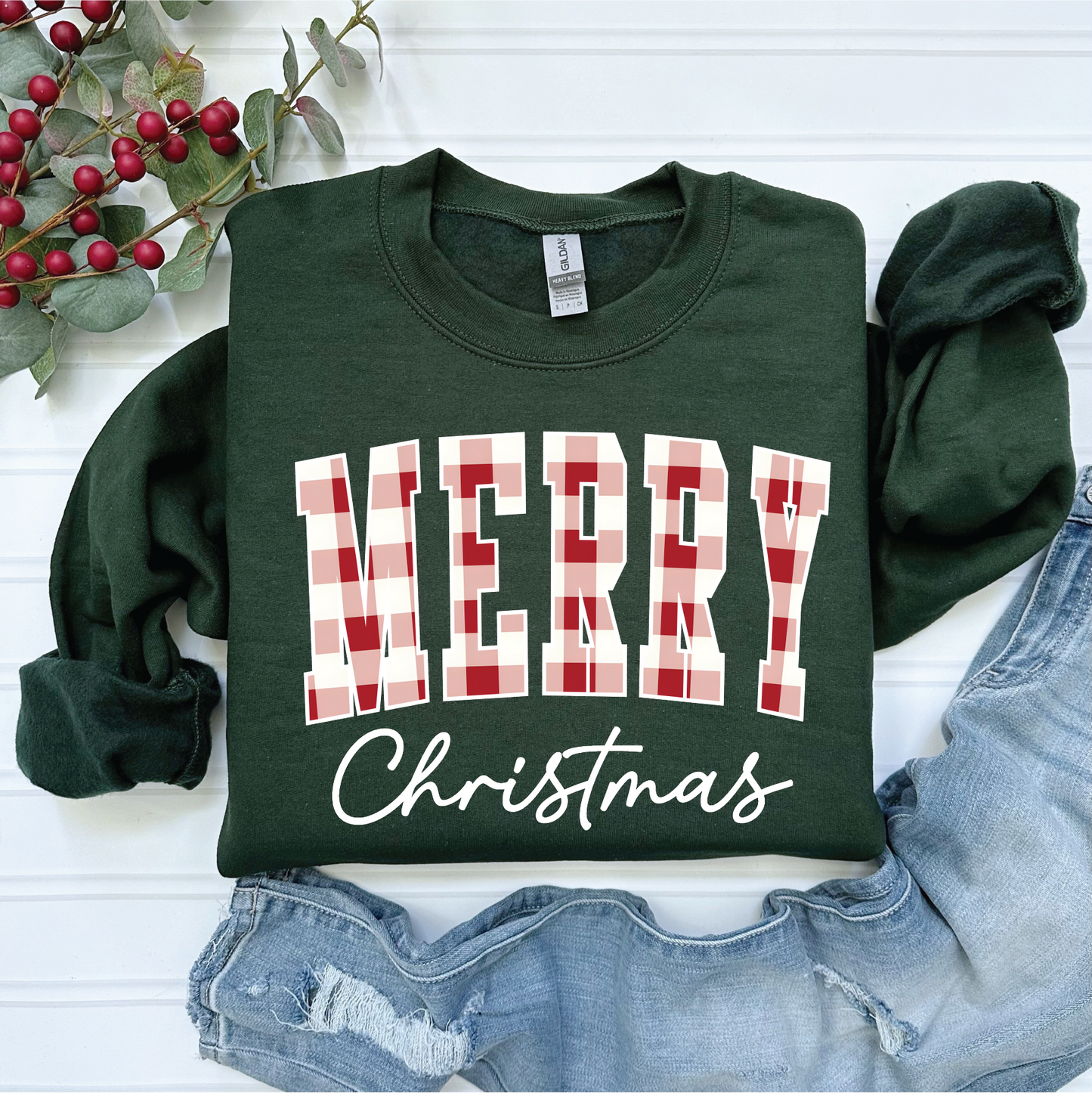 The Merry Christmas Plaid sweatshirt by Southern Attitude Designs Inc features green fabric with "MERRY Christmas" in plaid lettering, shown on a white surface alongside red berries and denim jeans.