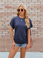 A woman in a Suhkasana Relaxed Fit Short Sleeve Rayon Blend Tee for women, navy blue, paired with denim shorts, sunglasses, and a brown shoulder bag stands before a brick wall.